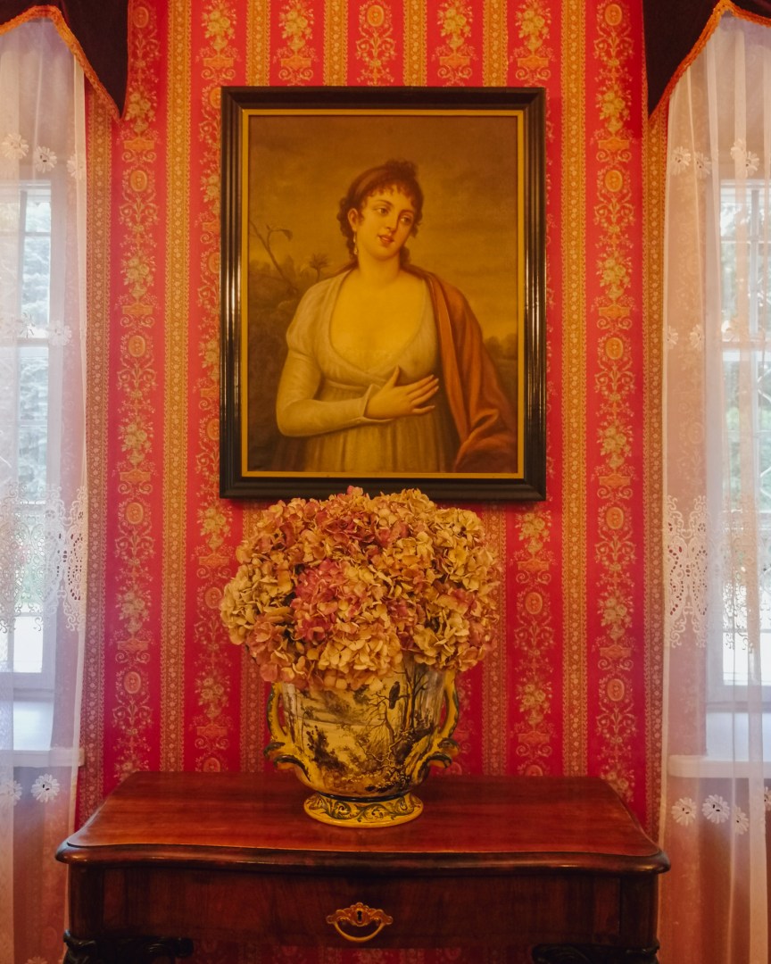 Oil portrait of a woman framed by red wallpaper and a flower arrangement.