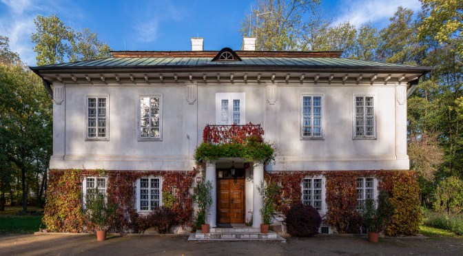 Front view of historic Polish manor house with white façade, ivy-covered walls, and decorative entrance