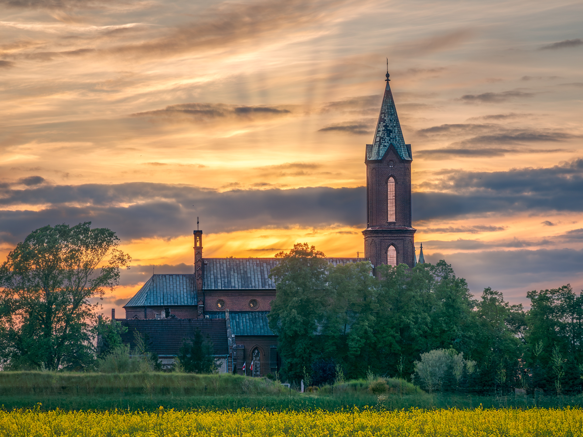 Sunset over the Church of the Immaculate Conception in Wojkow, Poland, with a blooming rapeseed field in the foreground and dramatic sky in the background
