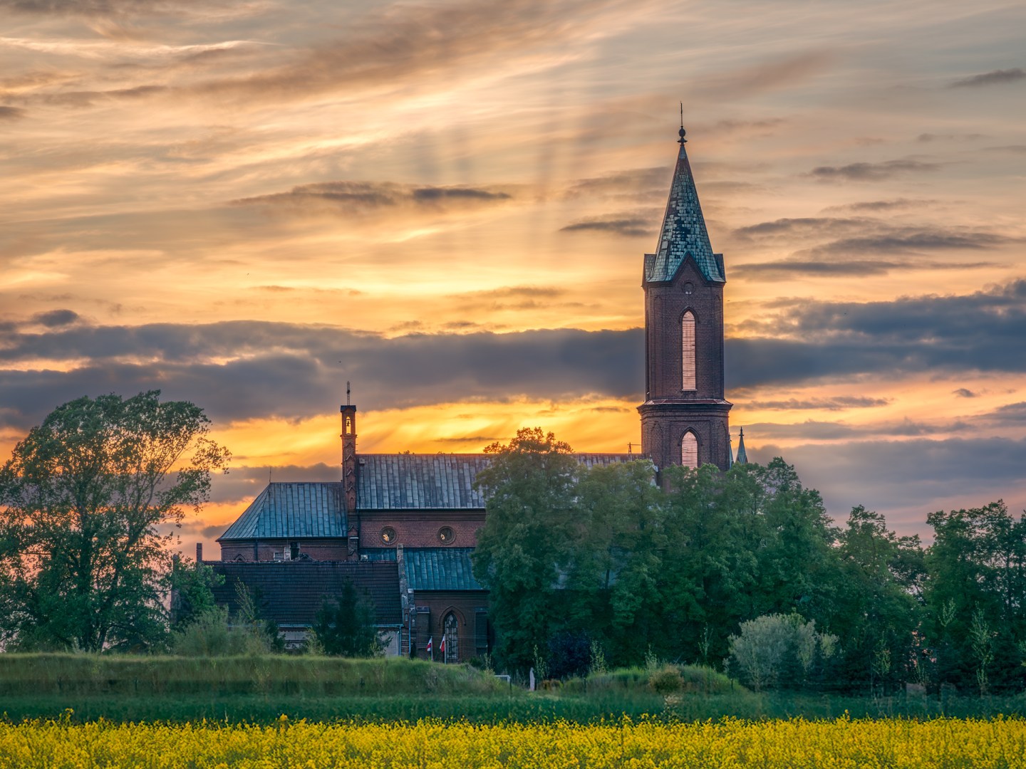 Sunset over the Church of the Immaculate Conception in Wojkow, Poland, with a blooming rapeseed field in the foreground and dramatic sky in the background