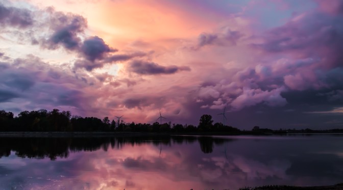 A serene sunset over a lake in western Poland with dramatic pink and purple clouds reflected in the water, and wind turbines silhouetted against the horizon