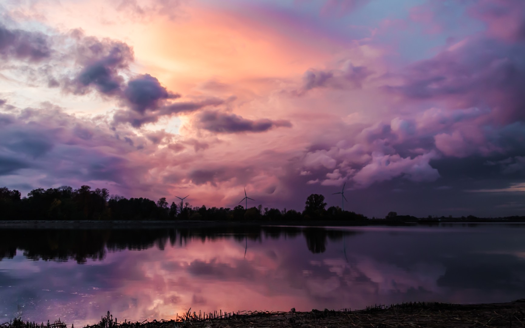 A serene sunset over a lake in western Poland with dramatic pink and purple clouds reflected in the water, and wind turbines silhouetted against the horizon