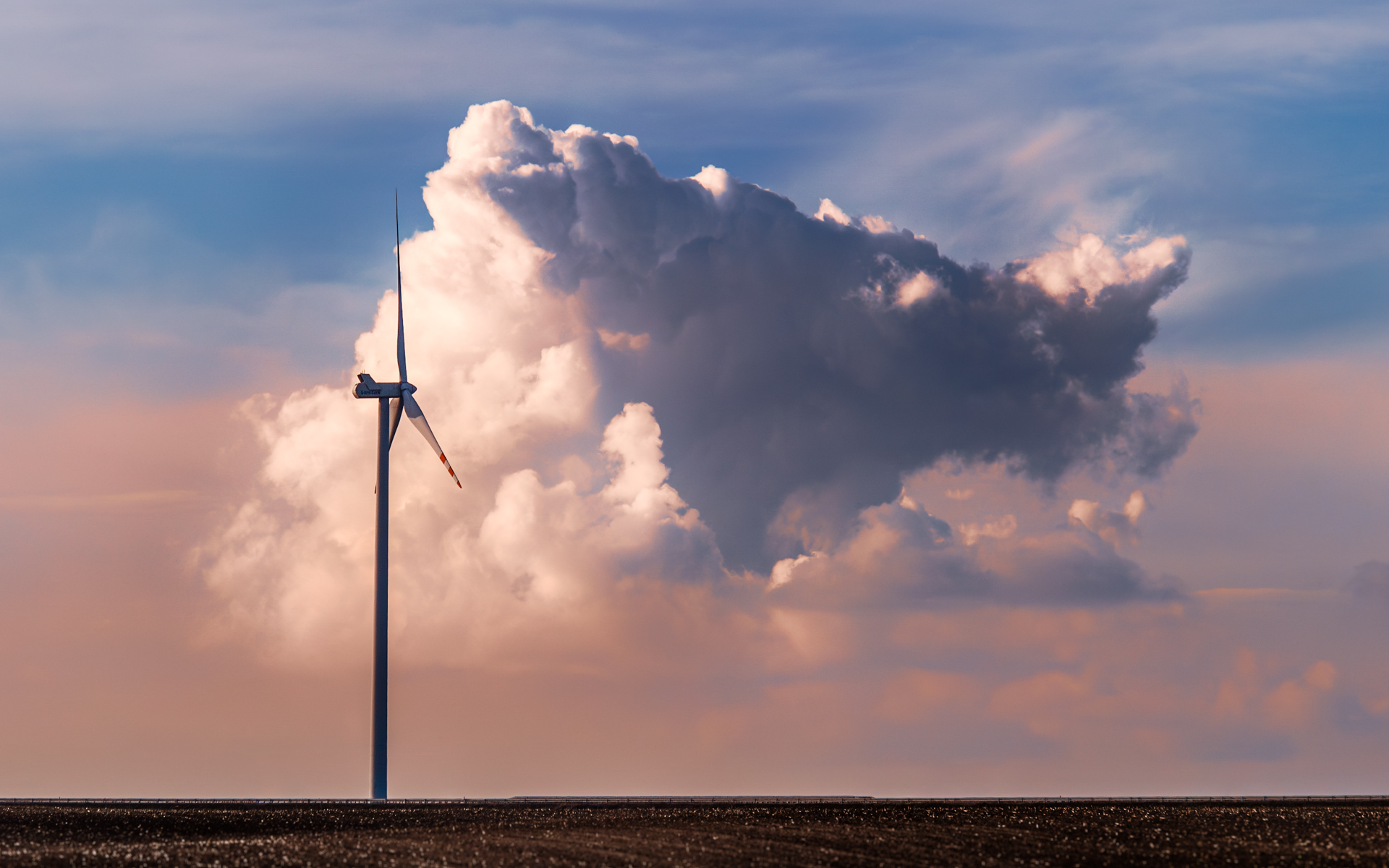 A single wind turbine stands against a dramatic cumulus cloud at sunset, with a flat dark field in the foreground and soft pastel tones in the sky