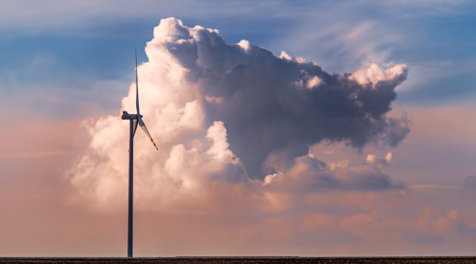 A single wind turbine stands against a dramatic cumulus cloud at sunset, with a flat dark field in the foreground and soft pastel tones in the sky