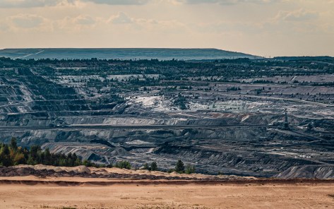 A wide view of the stepped layers of Bełchatów brown coal mine in Poland, with exposed soil, mining machinery, and distant forest under a hazy spring sky
