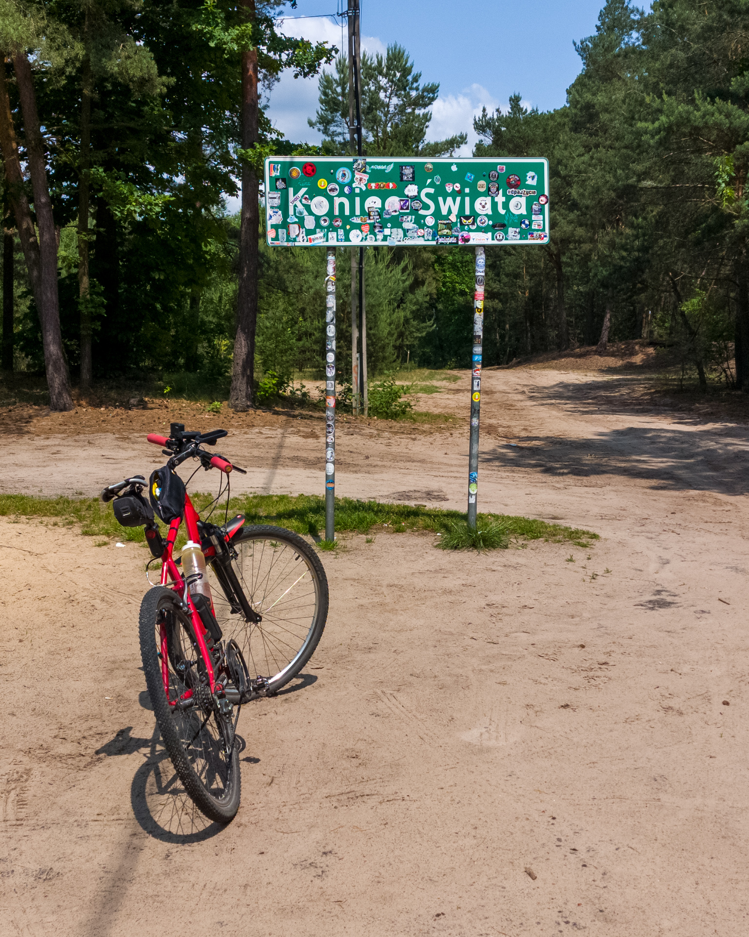 Red bicycle leaning on the sticker-covered Koniec Swiata sign at a forest dead-end in central Poland