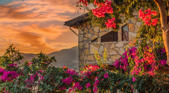 Rustic Mediterranean stone house surrounded by blooming bougainvillea, captured at sunset with vibrant skies and distant hills
