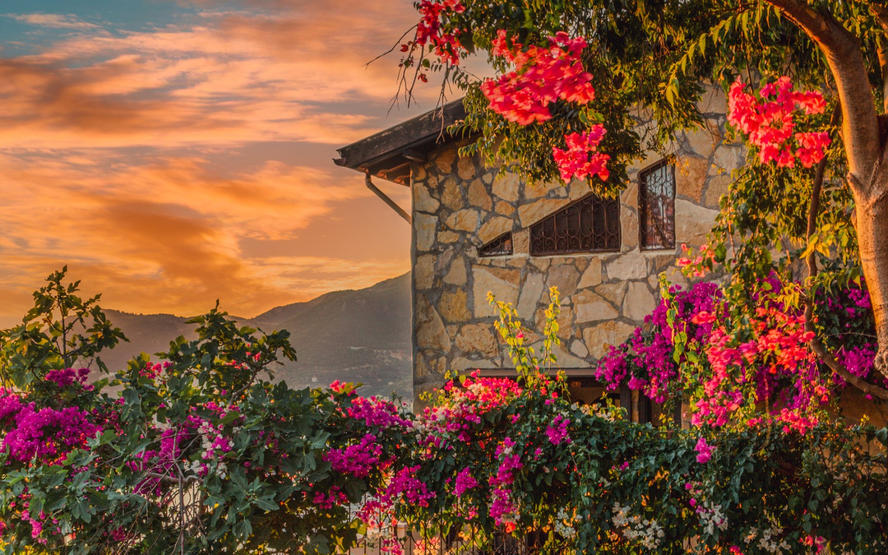 Rustic Mediterranean stone house surrounded by blooming bougainvillea, captured at sunset with vibrant skies and distant hills