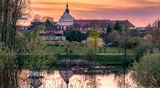 Sunset over the 13th-century cloister in Sieradz, Poland, with its reflection in a quiet pond surrounded by spring trees and greenery