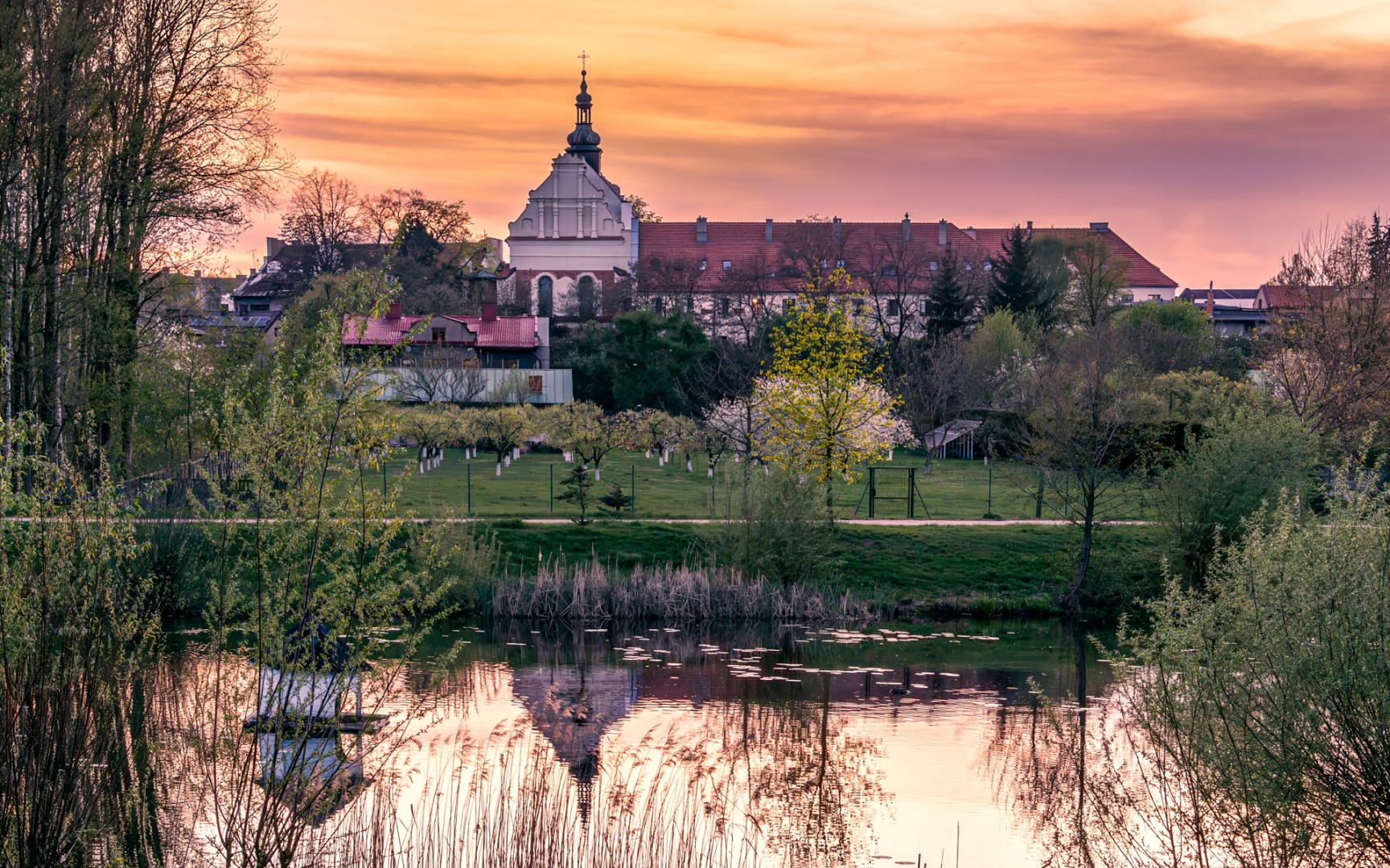Sunset over the 13th-century cloister in Sieradz, Poland, with its reflection in a quiet pond surrounded by spring trees and greenery
