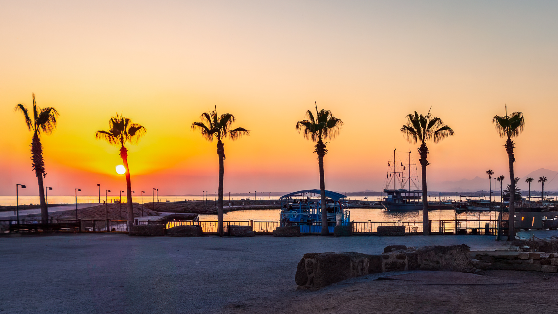Sunset over a Mediterranean harbor with palm trees silhouetted against a golden sky, moored boats in the water, and distant mountains on the horizon