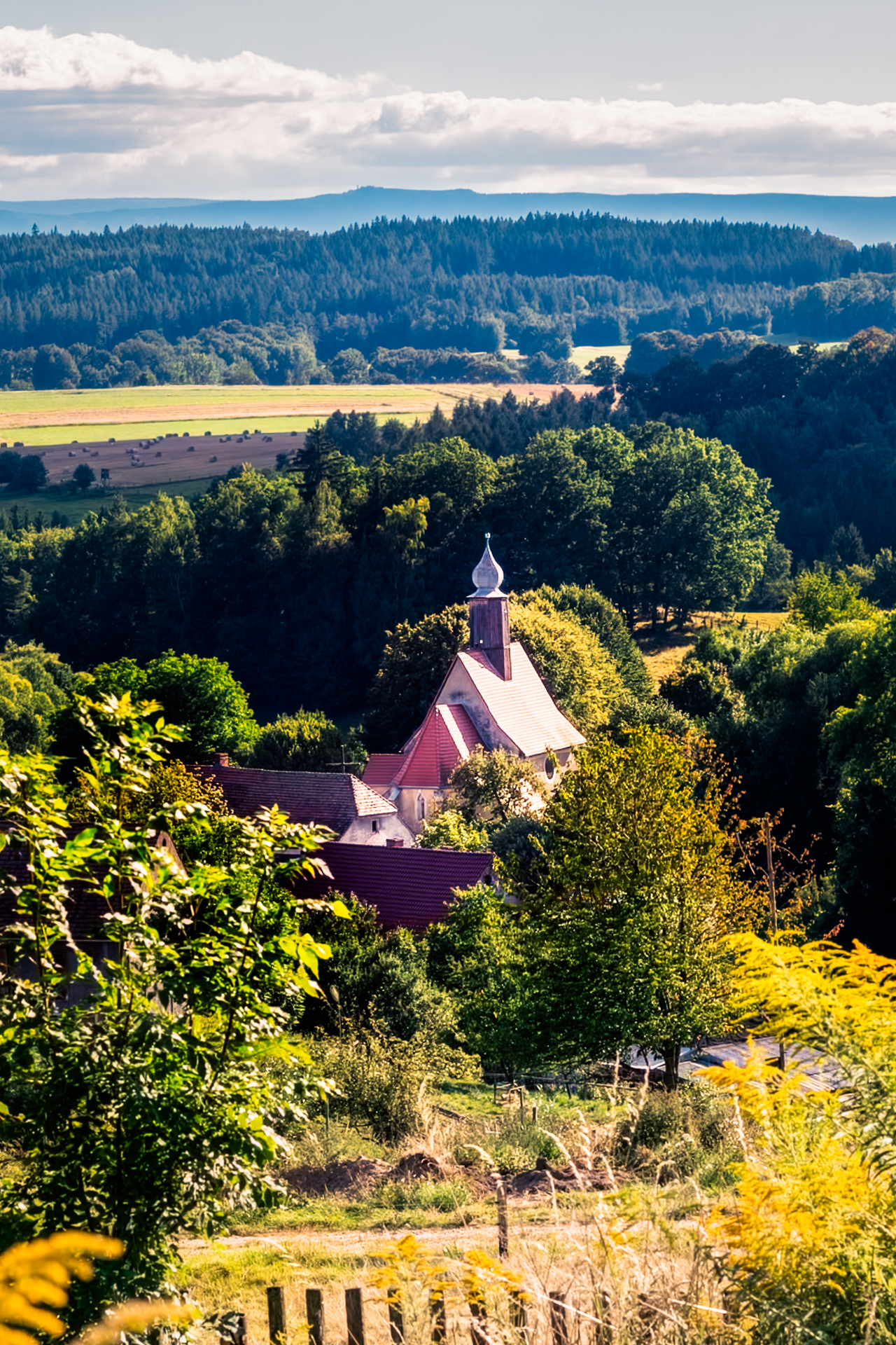 Countryside church with red roof among fields, meadows, and forest ridges on a sunny summer day