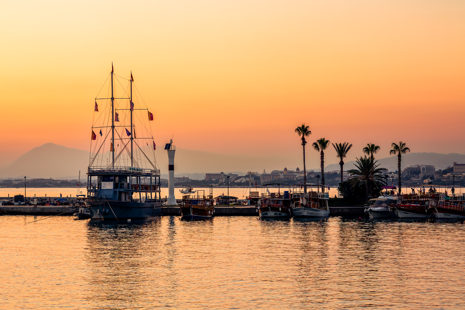 A serene sunset view over a Mediterranean harbor. A large ship with tall masts is moored on the left, balanced by palm tree silhouettes on the right. The sky is soft with shades of orange, pink, and gold, reflecting on the calm water