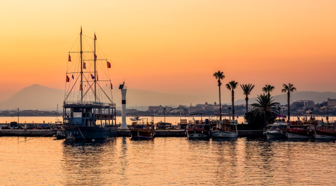 A serene sunset view over a Mediterranean harbor. A large ship with tall masts is moored on the left, balanced by palm tree silhouettes on the right. The sky is soft with shades of orange, pink, and gold, reflecting on the calm water