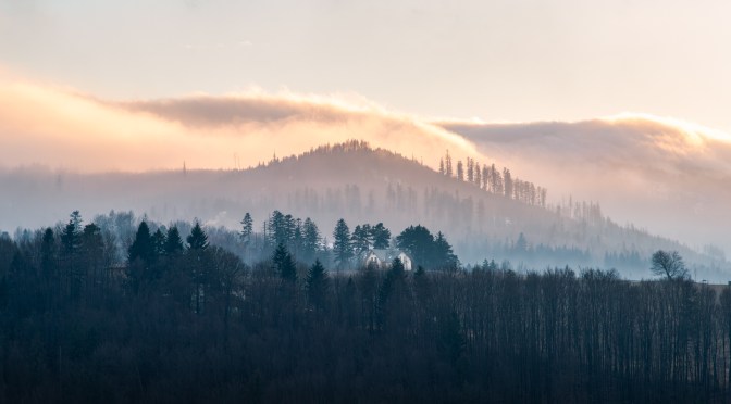 Winter landscape in Wisła with layered hills, soft morning fog, a small hut in the midground, and warm sunrise light above the ridge