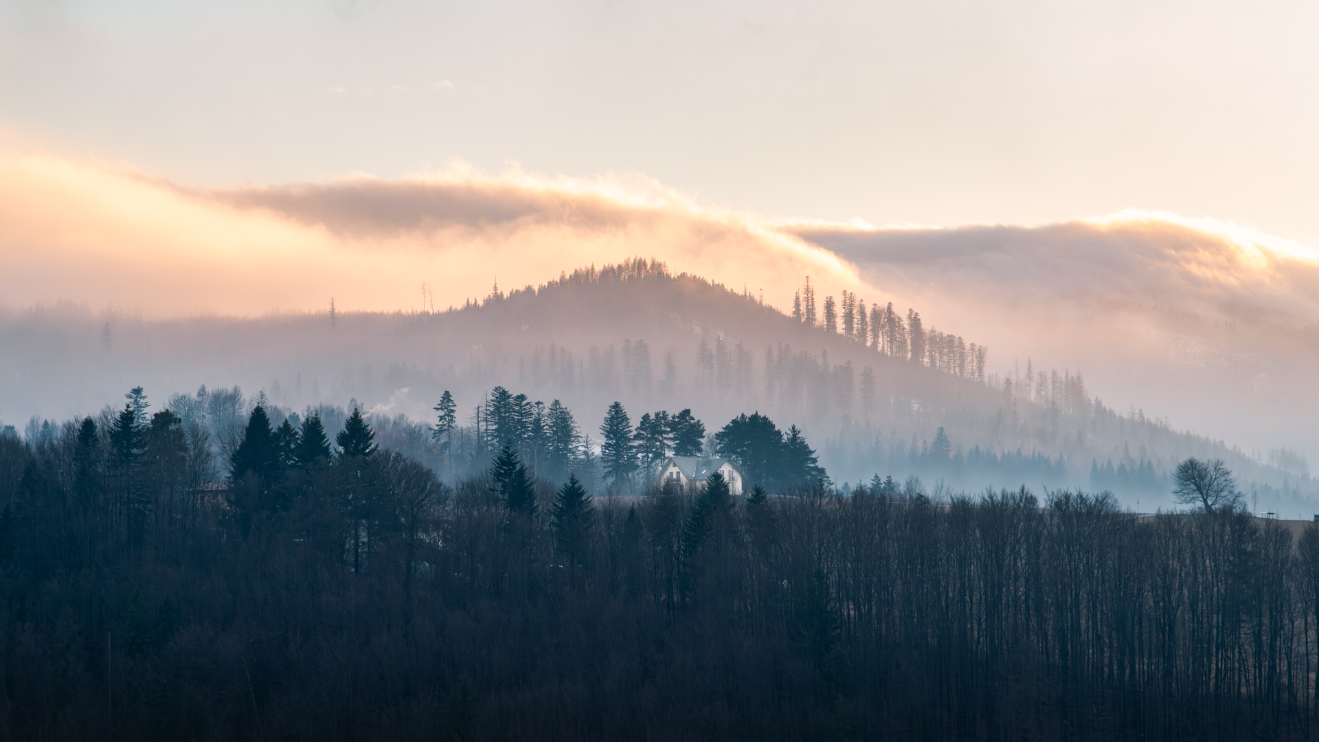 Winter landscape in Wisła with layered hills, soft morning fog, a small hut in the midground, and warm sunrise light above the ridge