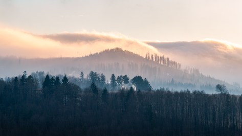 Winter landscape in Wisła with layered hills, soft morning fog, a small hut in the midground, and warm sunrise light above the ridge